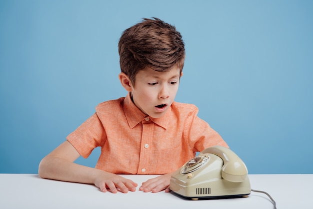 Ragazzino confuso con il vecchio telefono seduto al tavolo sfondo blu