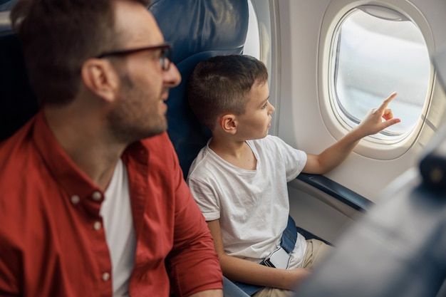 Ragazzino curioso che guarda fuori dalla finestra mentre viaggia in aereo insieme a suo padre. Famiglia, trasporto, concetto di vacanza