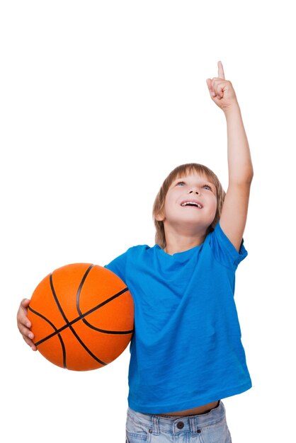 Ragazzino curioso. Ragazzino allegro che tiene la palla da basket e punta verso l'alto mentre sta in piedi isolato su bianco