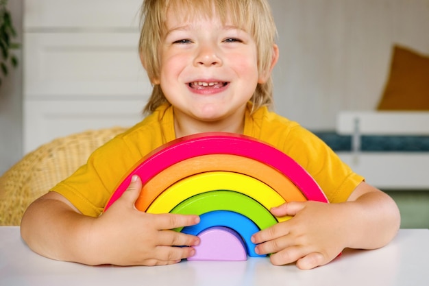 Ragazzino dai capelli biondi in vestiti di tessuto naturale gioca a giocattoli di legno color arcobaleno al tavolo bianco Eco