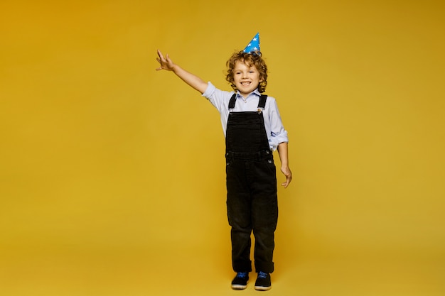 Ragazzino dai capelli rossi alla moda in tuta e protezione di compleanno in posa sullo sfondo giallo, isolato. Moda per bambini