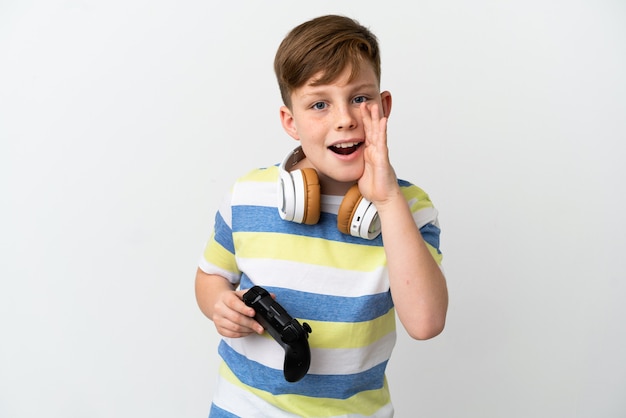 Ragazzino dai capelli rossi che tiene in mano un game pad isolato su una superficie bianca che grida con la bocca spalancata