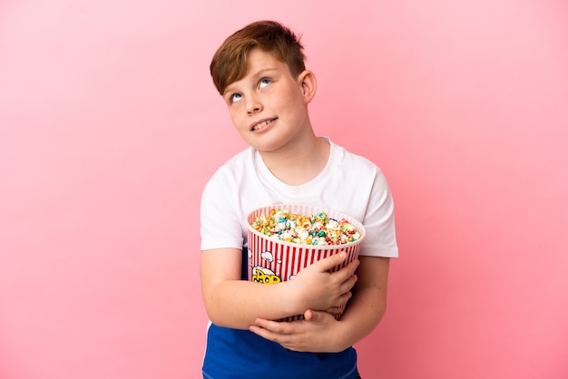 Ragazzino dai capelli rossi isolato su sfondo rosa con in mano un grosso secchio di popcorn