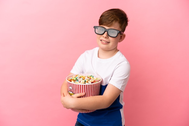 Ragazzino dai capelli rossi isolato su sfondo rosa con occhiali 3d e con in mano un grosso secchio di popcorn