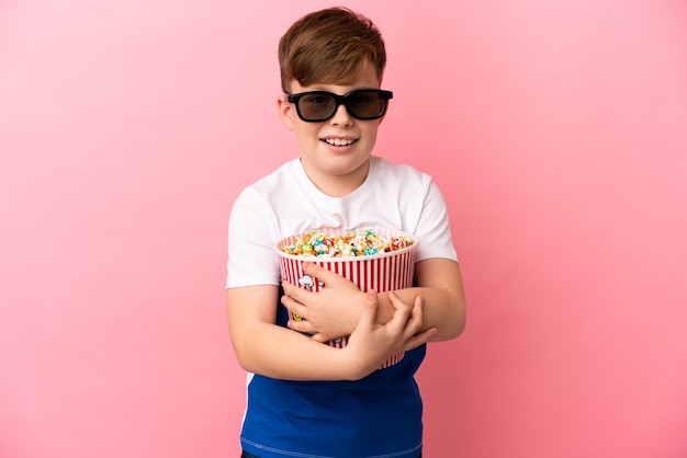 Ragazzino dai capelli rossi isolato su sfondo rosa con occhiali 3d e con in mano un grosso secchio di popcorn