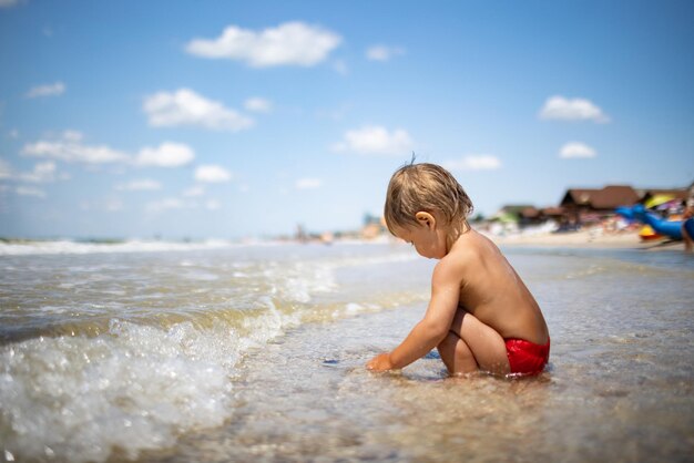 Ragazzino divertente e divertente, raccoglie conchiglie e ciottoli nel calmo mare blu su un fondo sabbioso sotto il caldo sole estivo in una vacanza luminosa