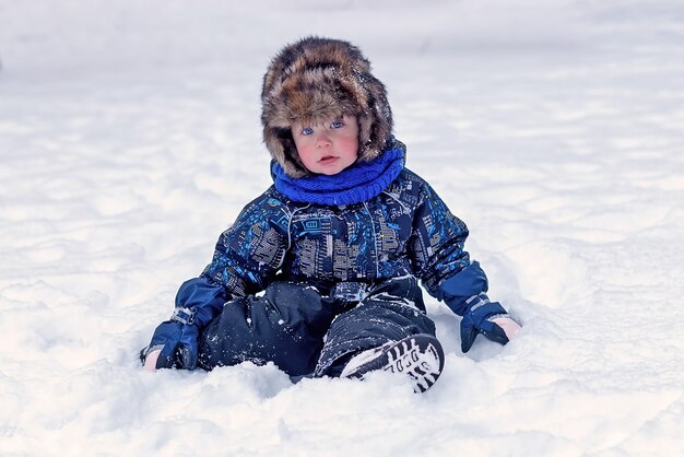 Ragazzino divertente in tuta e vestiti del cappello di pelliccia che giocano all'aperto durante la nevicata. Riposo attivo con i bambini in inverno nelle fredde giornate nevose. Bambino felice.