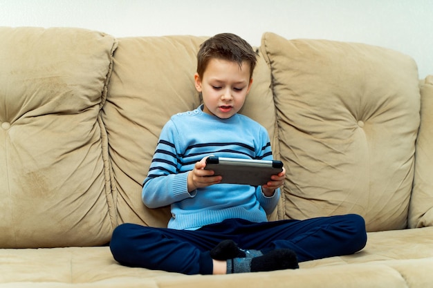 Ragazzino entusiasta in abiti casual seduto su un comodo divano e giocando su un tablet Ragazzo con tablet digitale nelle sue mani sul divano