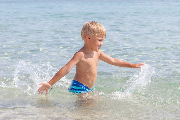 Ragazzino felice divertente sveglio che gioca nelle onde di acqua in mare oceano in una giornata di sole