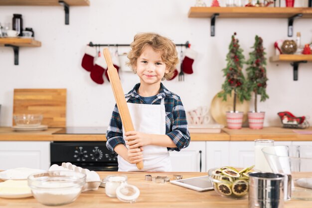 Ragazzino felice in grembiule che tiene il mattarello mentre fa i biscotti in cucina per aiutare sua madre