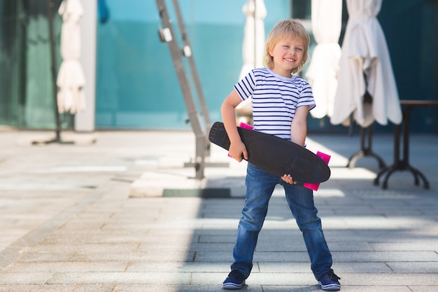 Ragazzino grazioso su una tavola da skate. Ragazzo emotivo all'aperto. Bambino carino pattinaggio indossando occhiali da sole.