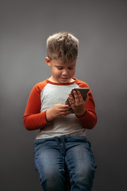 Ragazzino in camicia rossa che guarda lo smartphone tenendolo tra le mani e sorride isolato su grigio...