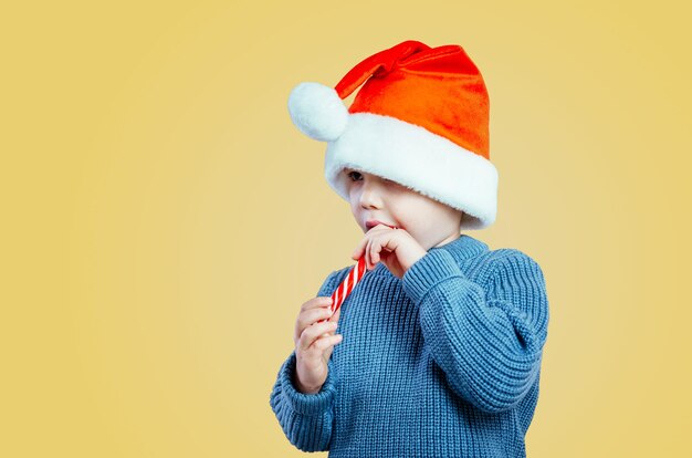 Ragazzino in cappello di Babbo Natale che mangia lecca-lecca di bastoncini di zucchero lo a Natale su sfondo giallo