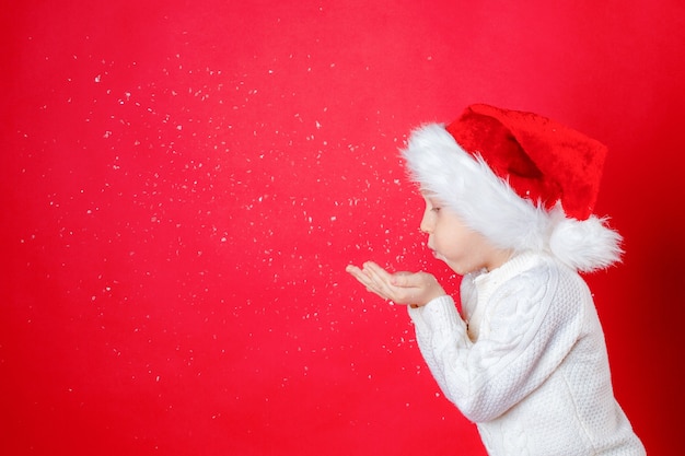 Ragazzino in cappello di Babbo Natale che soffia neve su sfondo rosso capodanno