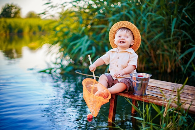 Ragazzino in cappello di paglia che si siede sul bordo di un bacino di legno e pesca nel lago al tramonto.