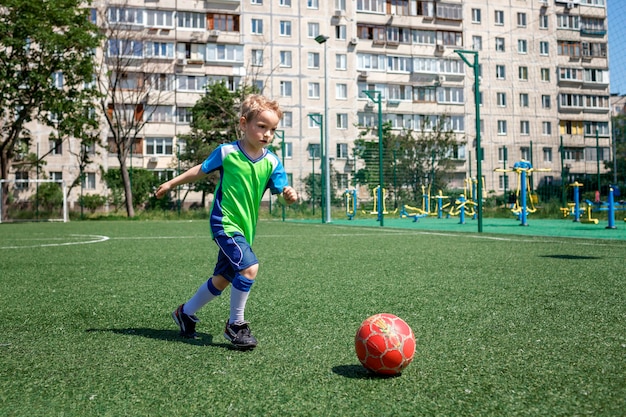 Ragazzino in forma blu e verde che gioca a calcio in campo aperto nel cortile un giovane calciatore