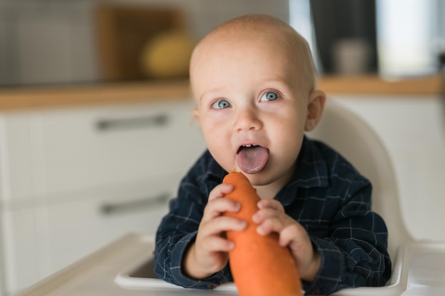 Ragazzino in una maglietta blu che si siede su una sedia per bambini che mangia lo spazio della copia della carota e spazio vuoto per t