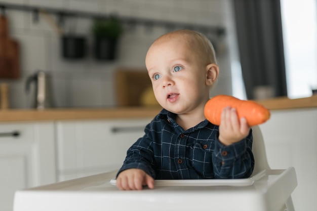 Ragazzino in una maglietta blu che si siede su una sedia per bambini che mangia lo spazio della copia della carota e spazio vuoto per t