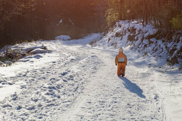 Ragazzino in una tuta arancione che cammina sulla strada innevata in una foresta di conifere. Giornata di sole invernale. Vista posteriore.