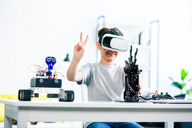 Ragazzino intelligente allegro seduto al tavolo e testando la sua creazione robotica per mezzo di tecnologie VR