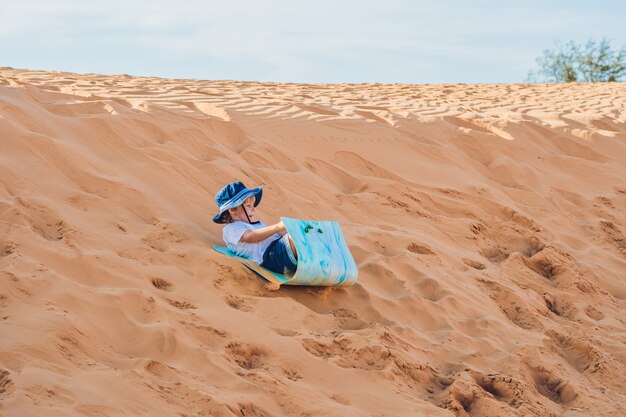 ragazzino sorridente felice con cappello rotola giù per le dune di sabbia nel deserto