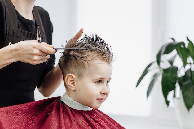 Ragazzino sorridente si fa tagliare i capelli con un paio di forbici da barbiere Primo piano di mani di donna che si toelettano i capelli di un ragazzo nel negozio di barbiere Ritratto di un bambino maschio dal negozio di barbiere per tagliarsi i capelli