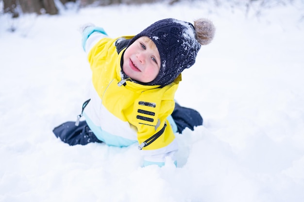 Ragazzino sorridente sveglio che si trova in un cumulo di neve, divertendosi, ridendo all'aperto nella foresta di inverno