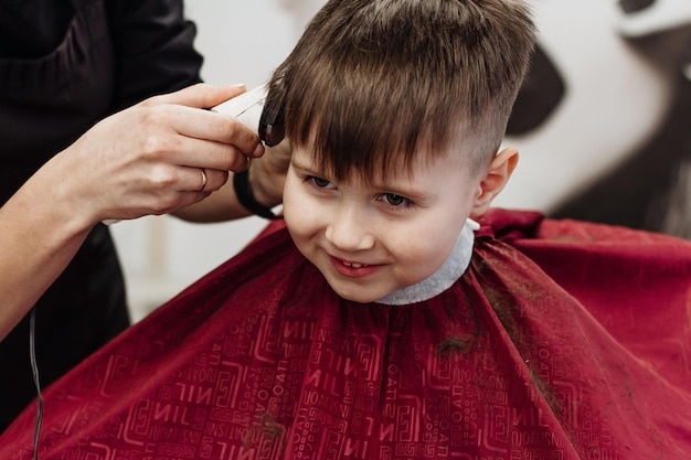 Ragazzino sorridente tagliato con la macchina del parrucchiere Primo piano delle mani della donna che toelettano i capelli del ragazzo del bambino nel negozio di barbiere Ritratto del bambino maschio dal negozio del barbiere per tagliarsi i capelli