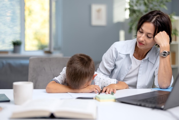 Ragazzino stanco che si appoggia la testa sulle mani mentre è seduto con la madre alla scrivania durante l'istruzione domestica Donna premurosa che aiuta suo figlio a fare i compiti