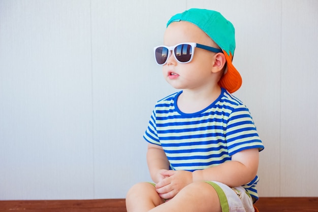 Ragazzino sveglio che indossa cappello e occhiali da sole su bianco