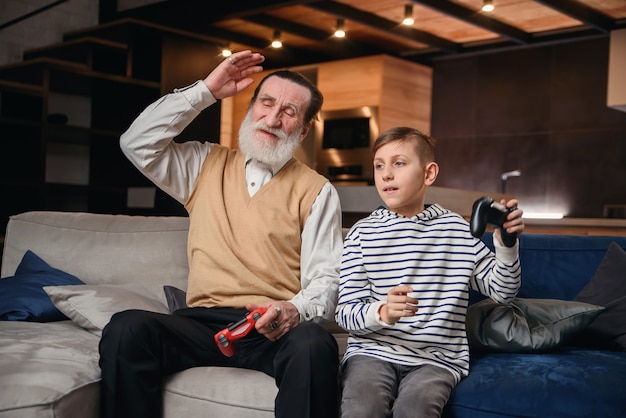 Ragazzino sveglio con il nonno che si siede sul sofà e che gioca video gioco con il game pad