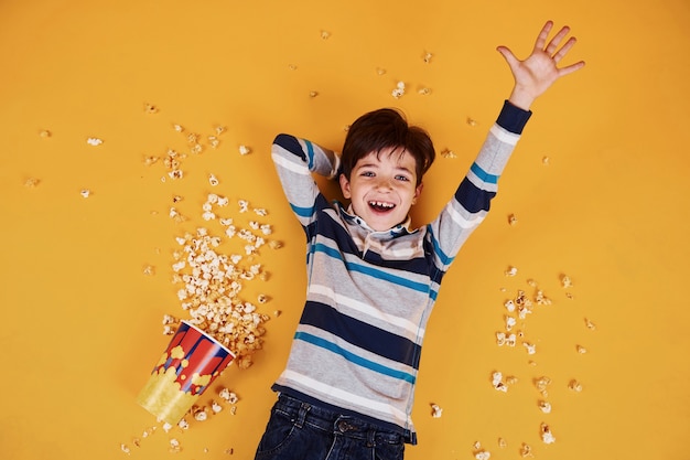 Ragazzino sveglio con popcorn sdraiato sul pavimento giallo.