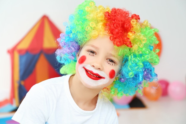 Ragazzino sveglio con trucco da clown in parrucca arcobaleno al chiuso. Festa dello sciocco di aprile