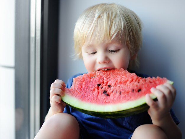 Ragazzino sveglio felice che mangia anguria che si siede sul davanzale. Cibo oragnico fresco per i più piccoli