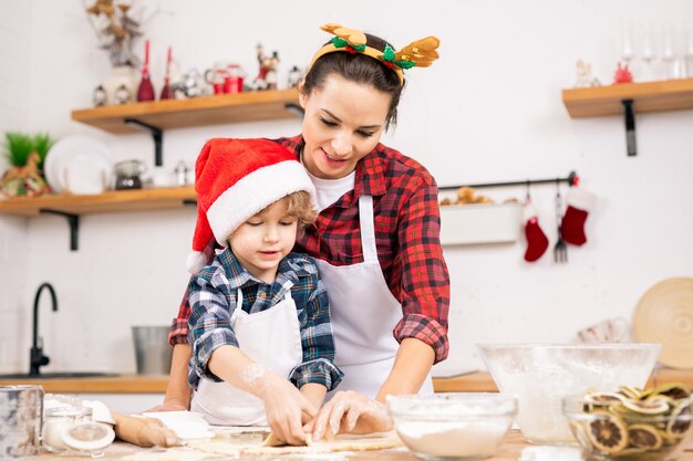 Ragazzino sveglio in protezione della Santa e sua madre nella fascia di Natale che fa i biscotti del pan di zenzero in cucina