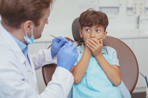 Ragazzino terrorizzato che copre la bocca con le mani, seduto su una sedia dentale