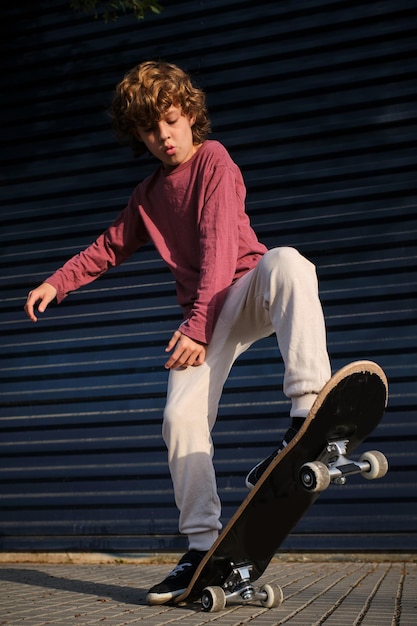 Ragazzo a tutta lunghezza che fa un trucco di skateboard sul marciapiede contro un muro blu scuro in una giornata di sole in città