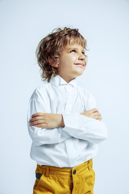 Ragazzo abbastanza giovane in abiti casual sul muro bianco. Posa alla moda. Bambino in età prescolare maschio caucasico con emozioni facciali luminose. Infanzia, espressione, divertimento. In posa mani incrociate.