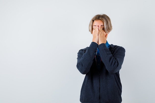 Ragazzo adolescente carino in camicia, felpa con cappuccio che copre il viso con le mani e sembra angosciato, vista frontale.