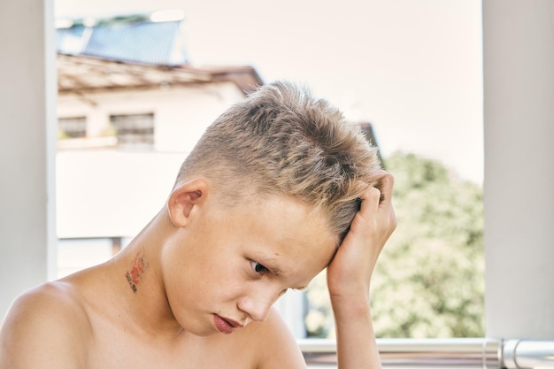 Ragazzo adolescente con capelli biondi corti puntelli testa con la mano seduta sul balcone dell'hotel