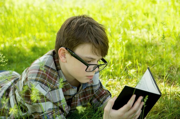 Ragazzo adolescente con gli occhiali sdraiato nell'erba verde e la lettura di un libro Vacanza all'aperto nel parco