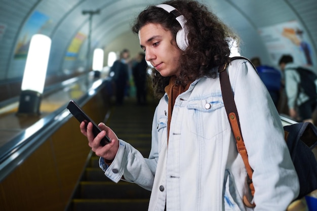 Ragazzo adolescente in abbigliamento casual e cuffie che utilizza lo smartphone mentre si trova sulla scala mobile