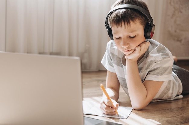 Ragazzo adolescente in cuffie wireless sdraiato sul pavimento con laptop con videochiamata e lezione online a casa Scolaro che studia i compiti Istruzione a distanza concetto di formazione a distanza