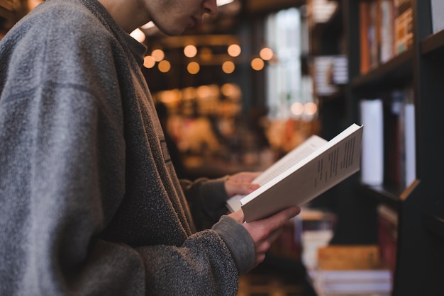 Ragazzo adolescente in possesso di un libro di carta in piedi nella biblioteca pubblica su scaffale e luce incandescente sullo sfondo Giovane uomo adulto che legge in negozio da vicino