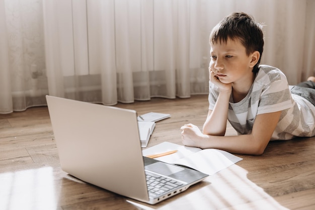 Ragazzo adolescente sdraiato sul pavimento con un laptop con videochiamata e lezione online a casa Scolaro che studia i compiti Istruzione a distanza Concetto di formazione a distanza