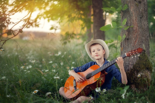 Ragazzo adorabile con la chitarra, rilassandosi nel parco. Scherzi la seduta su un'erba nel giorno di estate