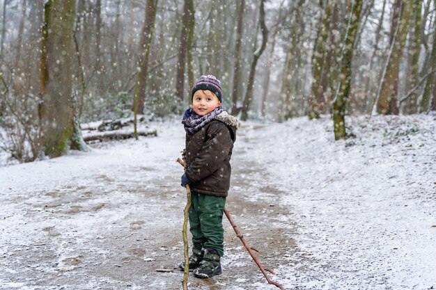 Ragazzo adorabile del bambino che cammina in un parco in inverno. Il bambino che indossa vestiti caldi gioca con bastoncini di legno in una foresta. Bambino che si gode la neve della prima volta.