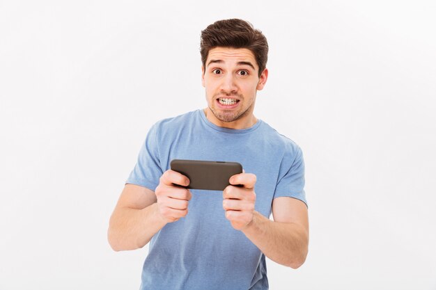Ragazzo adulto del gioco in maglietta casuale con tensione sul fronte che ha intrattenimento mobile mentre giocando sullo smartphone, isolato sopra la parete bianca