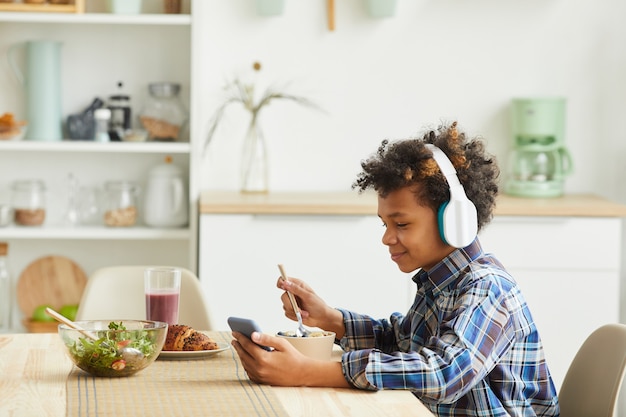 Ragazzo africano in cuffie utilizzando il suo telefono cellulare mentre si mangia la colazione in cucina