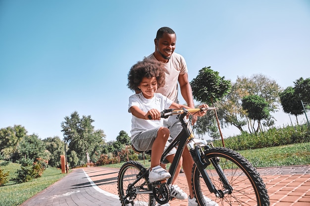 Ragazzo afroamericano concentrato che guarda i pedali mentre pedala accanto a suo padre